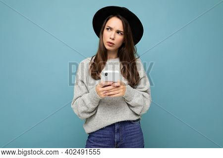 Beautiful Young Brunette Woman Thinking Wearing Black Hat And Grey Sweater Holding Smartphone Lookin