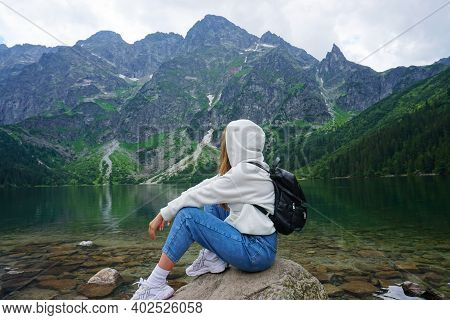 Girl In White Hoodie And Jeans With Backpack On His Back. Sits Sideways On A Rock In Front Of The La