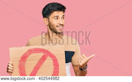 Young handsome man holding prohibited warning banner pointing thumb up to the side smiling happy with open mouth 