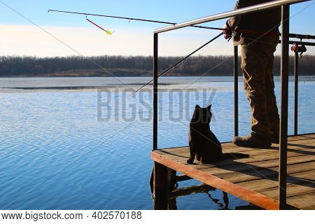 Black Cat Looks Back Sits Near The Silhouette Of A Fisherman, Fishing On The River, Blue Sky, Trees 