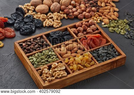 Various varieties of nuts, seeds and dried fruits lying in wooden box on black slate background. Top view. Healthy food. Vegetarian nutrition