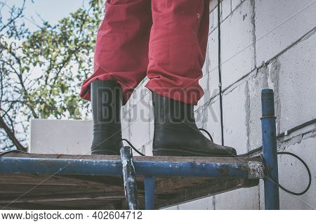 The Mans Legs Of A Builder In Rubber Boots Stand On The Scaffolding. Outdoor Construction Work, Past
