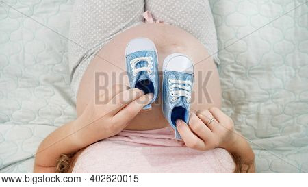 Top View Of Pregnant Woman With Big Belly Sitting On Bed And Playing With Little Newborn Baby Boots.