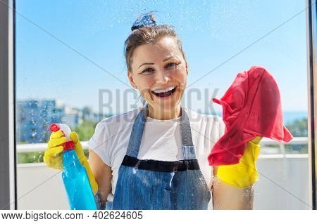 Portrait Of Mature Positive Woman Washing Window. Smiling Female In Gloves With Spray Bottle And Rag