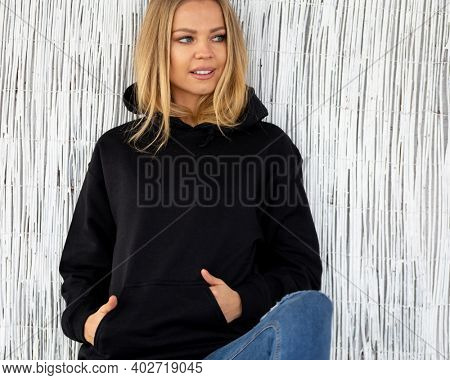 Sweatshirt hoodie mockup. A nice smiling blonde poses in a black sweatshirt against a white wall. She is sitting facing the camera.