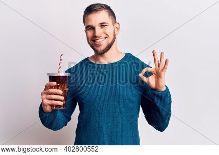 Young handsome man dirnking cola refreshment beverage doing ok sign with fingers, smiling friendly gesturing excellent symbol 