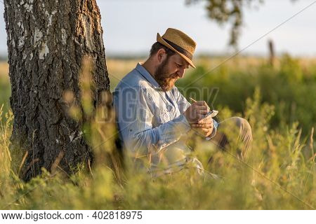 An Adult Man In A Hat With A Beard Writes Poetry Or Prose Sitting By A Tree. Nature Writing Concept