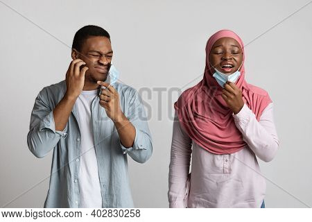 Irritated Muslim Black Couple Removing Face Masks To Breathe Over Grey Studio Background. African Ma