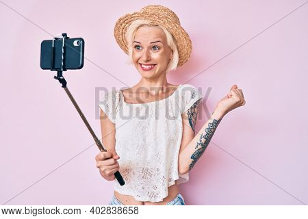 Young blonde woman with tattoo taking a selfie photo with smartphone screaming proud, celebrating victory and success very excited with raised arm 