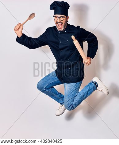 Middle age handsome hispanic man wearing glasses and cook uniform smiling happy. Jumping with smile on face holding kneader and spoon over isolated white background