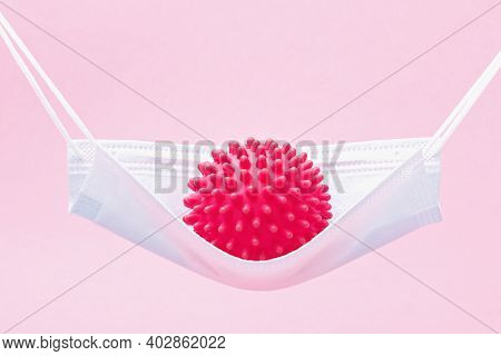 A Red Ball With Spikes Coronavirus Swinging In A Medical Mask Like In A Hammock Or Swing.