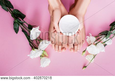 Top View Of Female Hands With Cosmetic Cream Near Feet And White Eustoma Flowers On Pink Background