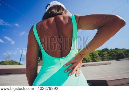 Back Pain. Athletic Woman In Pink Sportswear Standing At The Seaside Rubbing The Muscles Of Her Lowe