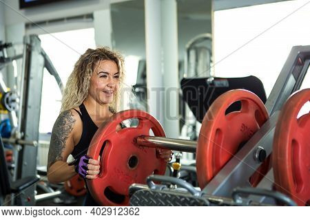 Woman With Long Curly Blonde Hair Holds A Heavy Barbell In His Hands. Bodybuilding, Trainer Of Fitne