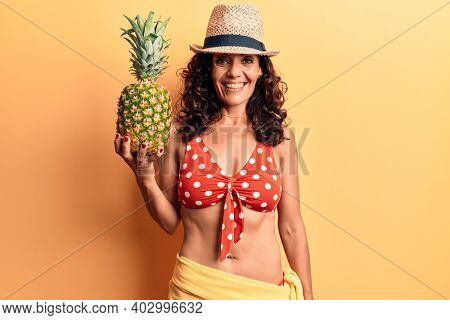 Middle age beautiful woman wearing bikini and summer hat holding pineappel looking positive and happy standing and smiling with a confident smile showing teeth 