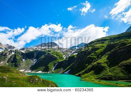 Turquoise Lake With Green Hills Around And The Snowy Mountains In The Background