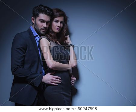 portrait of a young fashion couple looking into the camera while the man is holding the woman by her hips, from behind. on a dark blue background