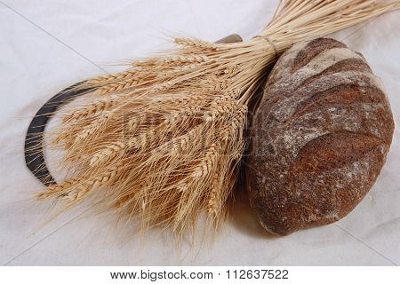 bunch of mown wheat ears with vintage handmade reaper hook sickle and rye dark french fresh bread loaf on white linen tablecloth