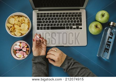 Work Space With Laptop, Candy, Chips, Water And Apple And Woman Hands On Blue Background. Top View