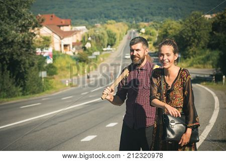 Couple of hikers happy smile on asphalt road on sunny day on natural landscape. Man with axe and woman with bag travel. Summer vacation active lifestyle traveling concept