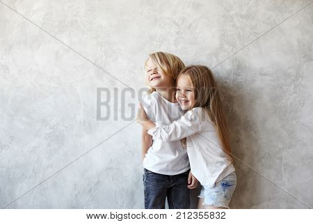 Joyful beautiful 5 year old female child with long straight hair hugging tight her elder brother both standing at blank wall and laughing. Puppy love between blonde boy and girl of preschool age
