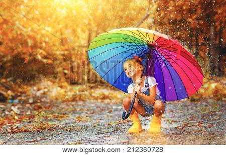 Happy funny ba child by girl with a multicolored umbrella jumping on puddles in rubber boots and laughing