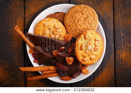 Oat and chocolate biscuits, black chocolate, nuts, raisins and a few cinnamon sticks on a plate on the table. Inside. View from above.