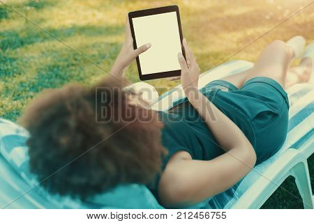 View from behind of curly female chilling on the lounge in park with mock-up of digital tablet with blank screen; rear view of woman resting on deck chair in the summer garden and using digital pad