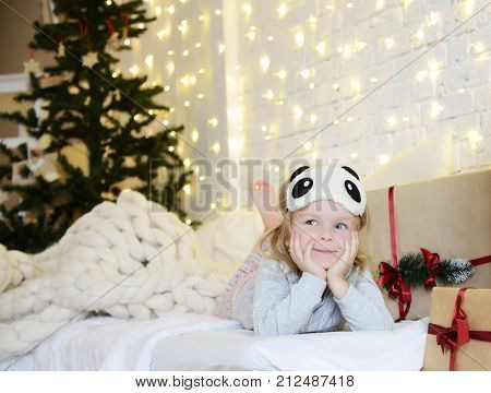 Young baby girl sitting dreaming near magical New year craft gifts by a Christmas tree on yellow magic lights background