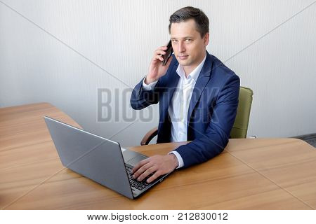 Handsome young concentrated attractive man using cellphone while working with laptop in the office.