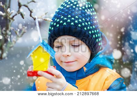 Little kid boy feeding birds in winter. Child hanging colorful selfmade bird house on tree on frosty cold day. Happy preschooler in colorful wam clothes. Selective focus on child and feeder with seeds
