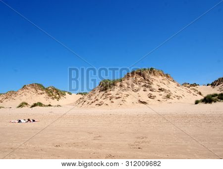 Sefton, Merseyside, United Kingdom - 27 June 2019: Two Women Sunbathing On The Beach With Grass On T