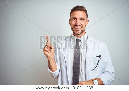 Young handsome doctor man wearing white profressional coat over isolated background with a big smile on face, pointing with hand and finger to the side looking at the camera.