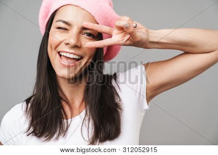 Image of a cute cheery young french woman in beret posing isolated over grey wall background showing peace gesture.