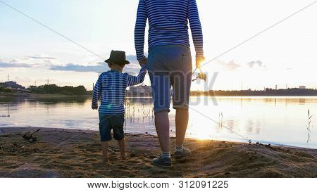 Father And Son In Striped Shirts With Fishing Rods Staying On The Riverside At Sunset