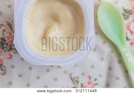 Baby Yogurt Cup With Creamy Apricot Yoghurt On Printed Table Cloth With Green Baby Spoon - Overhead 