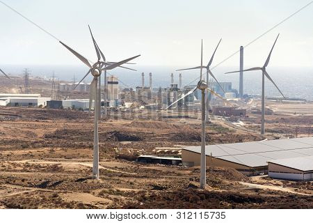 White Turbines At Wind Farm With Power Station And Sea In Background, Arid Landscape On Sunny Summer