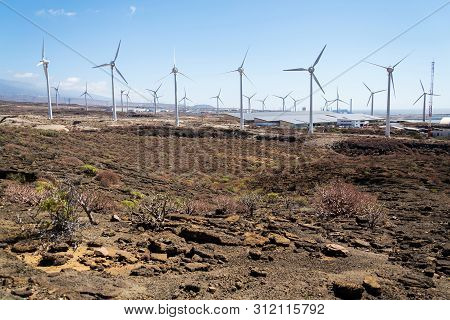 White Turbines At Wind Farm With Power Station And Sea In Background, Arid Landscape On Sunny Summer