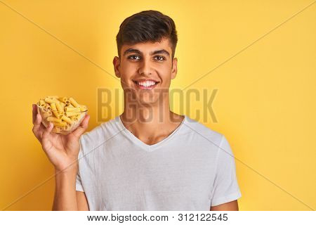 Young indian man holding bowl with dry pasta standing over isolated yellow background with a happy face standing and smiling with a confident smile showing teeth