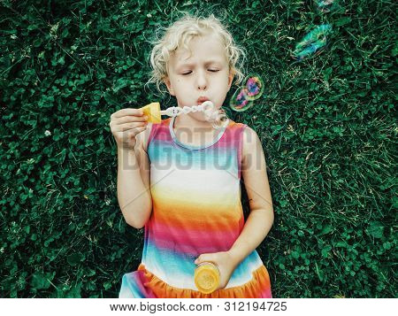 Candid Portrait Of Cute Funny Little Blonde Caucasian Child Girl Lying On Ground Grass Blowing Soap 