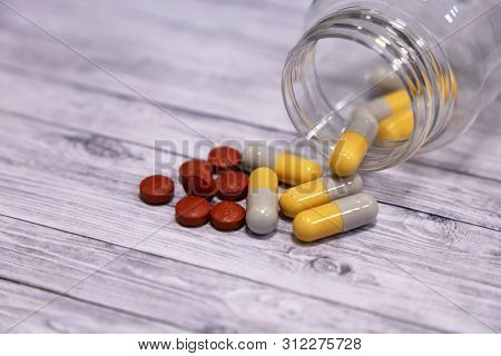 Pills And Capsules Spilled From Transparent Bottle On Vintage Light Wooden Table. Concept Of Drugs, 