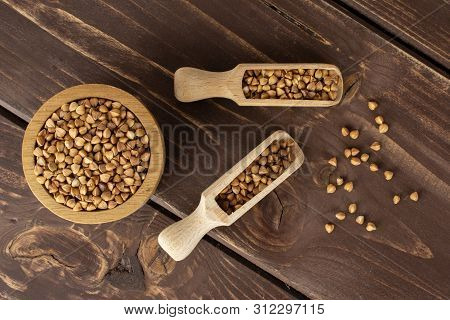 Lot Of Whole Raw Brown Buckwheat Grain In A Wooden Bowl With Wooden Scoop Flatlay On Brown Wood