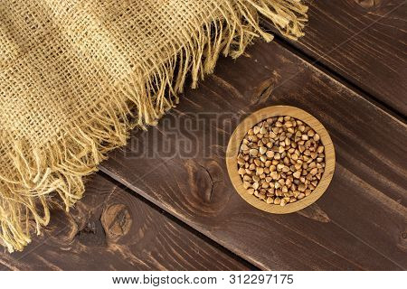 Lot Of Whole Raw Brown Buckwheat Grains In A Wooden Bowl With A Piece Of Jute Cloth Flatlay On Brown