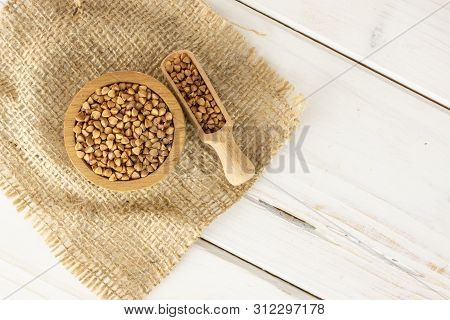 Lot Of Whole Raw Brown Buckwheat Grain In A Wooden Bowl On Jute Cloth With Wooden Scoop Flatlay On W