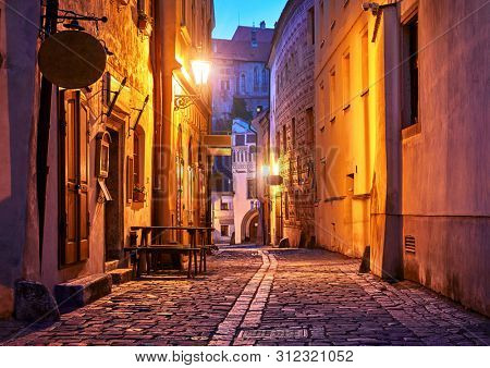 Narrow street of Old town with Night illumination. Czech Krumlov. Czech Republic. Shining lanterns at walls of medieval houses and road of paving stones.