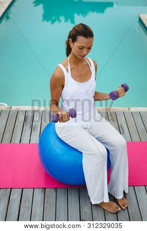 High angle view of fit mixed-race woman exercising with dumbbells while sitting on a exercise ball near swimming pool in the backyard. Summer fun at home by the swimming pool