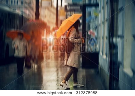 While Passers Go By, A Tourist Girl With An Orange Umbrella Stands On The Street Near The Store, And
