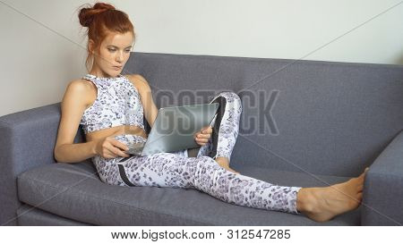 Young Fitness Woman In Sportswear Doing Yoga Stretching Leg While Using Laptop Or Computer Notebook 