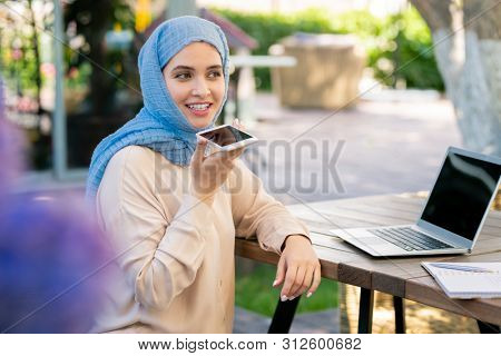 Happy girl in hijab recording voice message on smartphone in park