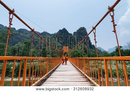 Vang Vieng, Laos - April 2019: Two Buddhist Monks In Orange Robes Crossing Suspension Bridge.